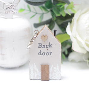 A small Chunky House Wooden Keyring with the words "Back door" written on it. The keyring features a wooden door, a heart-shaped accent, and a small nail on its roof. Placed on a white surface with blurred green foliage and flowers in the background, it makes for charming home decor.