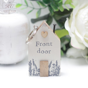 A charming, house-shaped keyholder with the words "Front door" printed on it. This piece of home decor features a heart and floral design and includes a Chunky House Wooden Keyring. In the background, white flowers and a white container enhance its appeal.