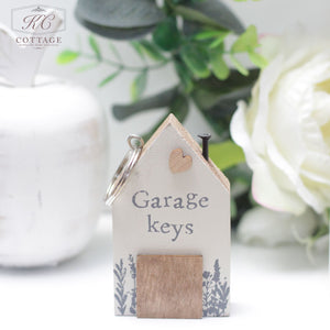 A small Chunky House Wooden Keyring in the shape of a house with the words "Garage keys" printed on it. Made of wood and painted white, it's adorned with a small wooden heart and a floral design at the bottom. Perfect for home decor, it sits in front of a white vase and flower in the background.