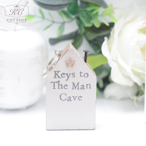 A small Chunky House Wooden Keyring shaped like a house with a star decoration at the top left is perfect for home decor. It has the text "Keys to The Man Cave" written on it. A keyring and nail are attached for hanging keys, set against a background of greenery and a faintly visible white vase.