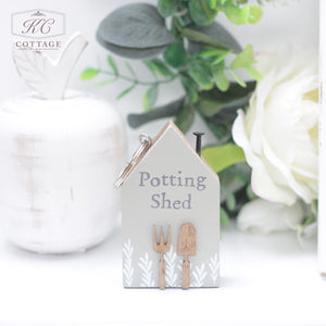 A small, house-shaped keychain reads "Potting Shed" and features illustrations of a shovel and trowel. Placed in front of green foliage with a white decorative object, the background includes a white flower. This charming Chunky House Wooden Keyring adds a touch of home decor to any setting.