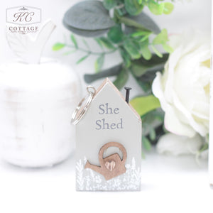 A small decorative key holder shaped like a house has the words "She Shed" written on it, complemented by a watering can illustration. It's adorned with a metal keyring and placed amid green leaves and white flowers, making it a charming piece of home decor. The **Chunky House Wooden Keyring** adds an extra touch of rustic charm to the setting.