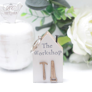 A small, house-shaped key holder with the text "The Workshop" on it, featuring miniature wooden hammer and saw decorations. Perfect for home decor, it's placed on a white surface with a white vase and greenery in the background. The Chunky House Wooden Keyring is attached at the top.