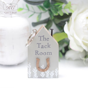 A decorative keychain shaped like a small house with "The Tack Room" text and a horseshoe on it. The Chunky House Wooden Keyring has a rustic design with white leaf patterns at the bottom, making it perfect for home decor. It is displayed with a white flower arrangement and greenery in the background.