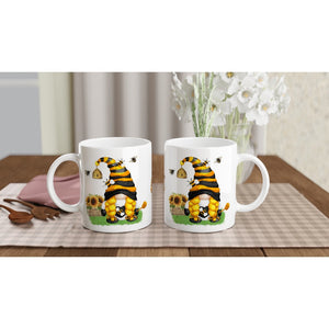 Two Bee & Sunflower Gonk Mugs on a dining table feature a cute cat wearing a bee costume, complete with sunflowers and little bees in the background. The table, adorned with a checkered tablecloth, has home decor accents like a plant and cutlery placed nearby.