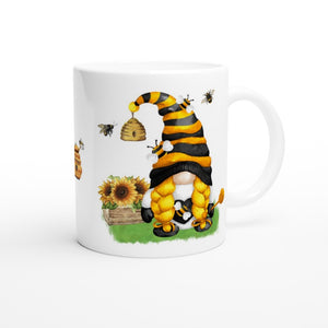 A charming white mug perfect for home decor features an illustration of a cute gnome with braided yellow hair, wearing a black and yellow striped hat. The gnome stands next to a beehive and sunflowers while several bees buzz around. The background is white, adding to its cheerful appeal. The product name is: Bee & Sunflower Gonk Mug.