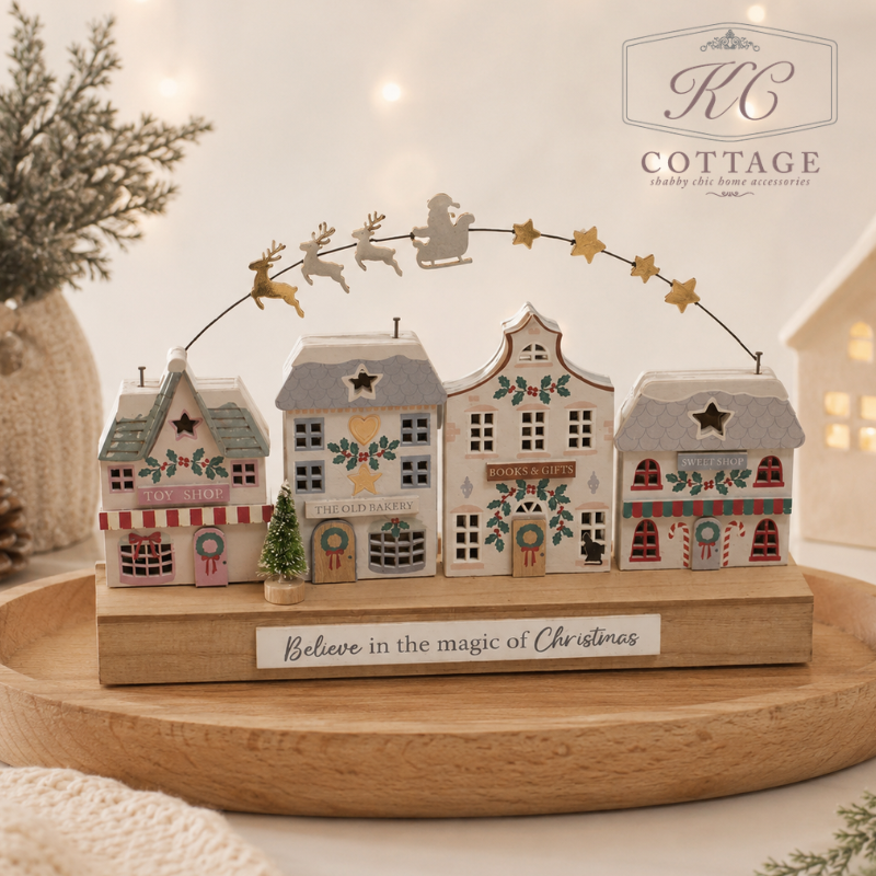 Christmas Home Decor Image. A decorative Christmas village tabletop display by KC Cottage, featuring four small painted houses on a wooden base with a curved wire of stars and Santa riding reindeer above. The display sits on a round wooden tray with a softly blurred warm holiday background.