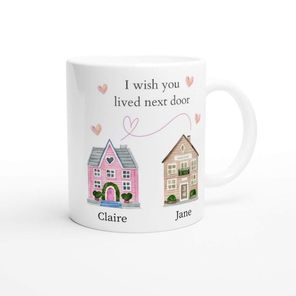 A white I Wish You Lived Next Door Personalised Mug showcases two illustrated houses connected by a pink heart-shaped line with the text "I wish you lived next door" above them. The pink house on the left is labeled "Claire," while the beige house on the right is labeled "Jane." Small pink hearts adorn this charming piece of home decor.