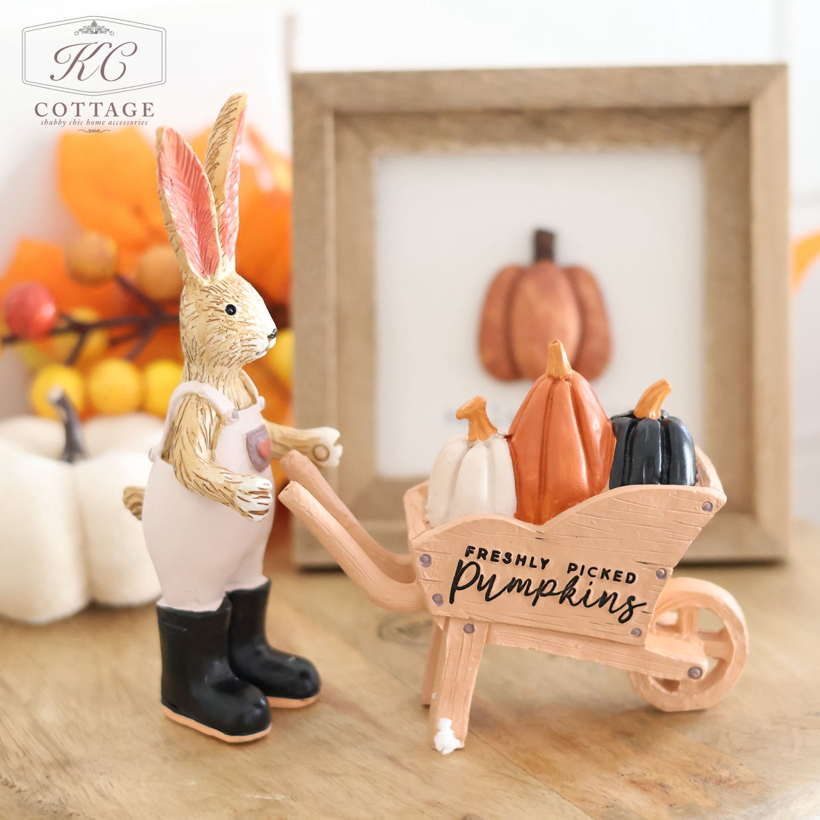 The Autumn Freshly Picked Pumpkins Wheelbarrow Rabbit, dressed in overalls and black boots, pushes a wooden wheelbarrow brimming with colorful pumpkins. Behind the ornament are autumn-themed decorations and a framed pumpkin illustration. The wheelbarrow features a sign that reads "Freshly Picked Pumpkins.