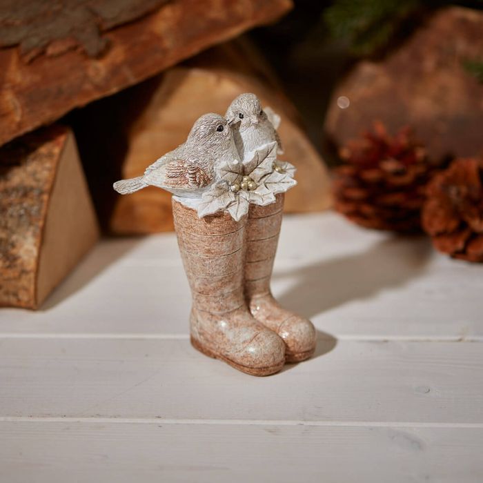 A decorative Holiday Decor figurine featuring two small birds perched on a pair of boots, the Christmas Birds Sitting On Wellies Ornament stands on a white wooden surface. It is surrounded by rustic elements, including wooden logs and a pine cone in the background.