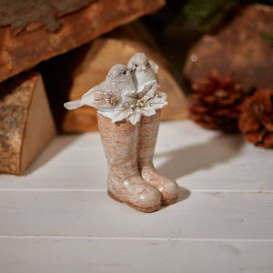 A decorative Holiday Decor figurine featuring two small birds perched on a pair of boots, the Christmas Birds Sitting On Wellies Ornament stands on a white wooden surface. It is surrounded by rustic elements, including wooden logs and a pine cone in the background.