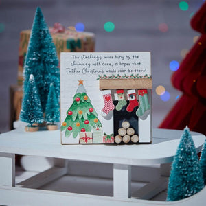 Introducing the Christmas 3D Fireplace Stockings Wooden Sign, a delightful festive decoration featuring a charming small house adorned with stockings hanging over a 3D fireplace. Inside, you'll find tiny logs, and beside it stands a decorated Christmas tree complete with ornaments and presents. This enchanting scene includes a wooden sign bearing a holiday message and is surrounded by miniature Christmas trees for an extra touch of seasonal cheer.
