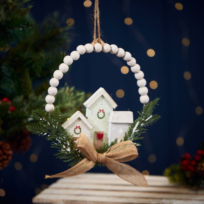 The Christmas Hanging Houses with Bead Circle Foliage & Bow showcases two small, white wooden houses adorned with green wreaths, nestled amid vibrant green pine branches. This charming ornament is encircled by a Wooden Beaded Circle and finished with a brown burlap bow, all suspended by a rustic string against a dark background.