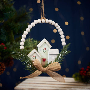 The Christmas Hanging Houses with Bead Circle Foliage & Bow showcases two small, white wooden houses adorned with green wreaths, nestled amid vibrant green pine branches. This charming ornament is encircled by a Wooden Beaded Circle and finished with a brown burlap bow, all suspended by a rustic string against a dark background.