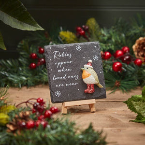 The Christmas Slate Robins Appear Easel Plaque features a small, decorative design surrounded by festive Christmas greenery and berries. It showcases a cute robin adorned with a hat and scarf, accompanied by the heartfelt text, "Robins appear when loved ones are near," set against a dark background embellished with snowflakes. This charming piece brings holiday cheer to any space.