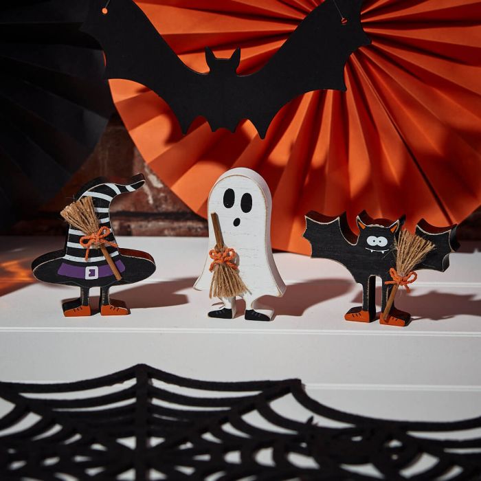 Three Halloween Wooden Standing Characters With 3D Broom stand atop a white shelf. From left to right: a witch with a pointy hat, a ghost, and a bat holding a 3D broomstick. In the background, orange and black paper decor resembling a fan and bat, as well as a black spider web, enhance the spooky ambiance of this charming Halloween decor.
