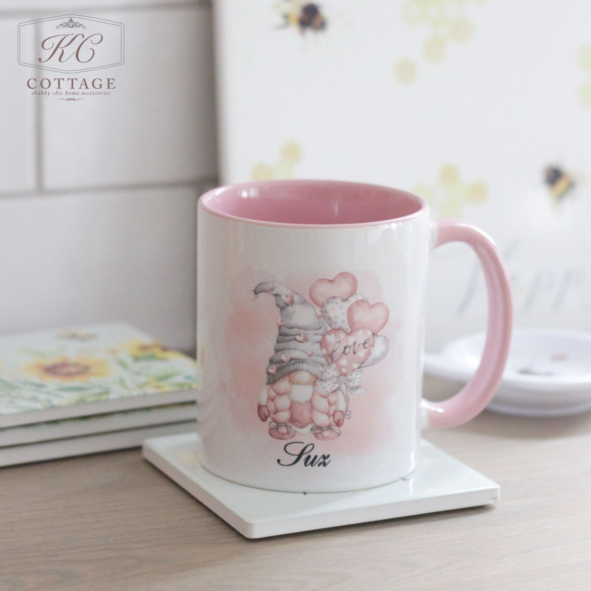 A Personalised Pink Love Gonk Mug sits on a square white coaster. The mug features an illustration of a gnome holding pink heart-shaped balloons with the word "Love" on one balloon. The name "Suz" is printed at the bottom, making it a charming piece of home decor.