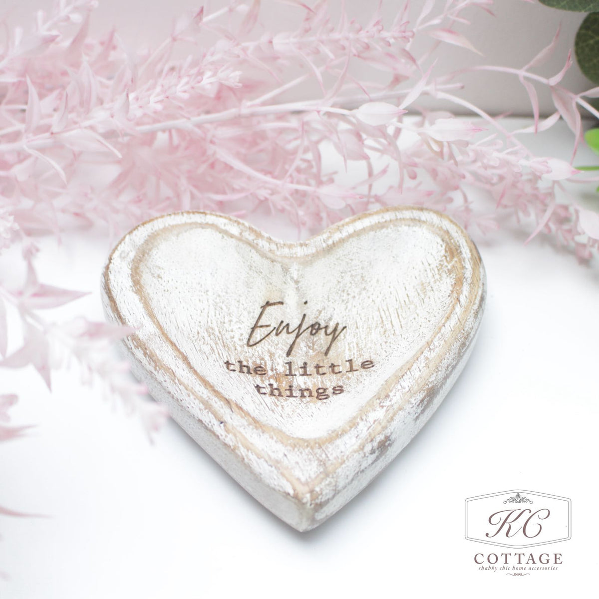 A rustic Mango Wooden Heart Trays with Sentiments piece with the words "Enjoy the little things" inscribed on it. The heart is set against a soft background with delicate pink foliage in the corner.