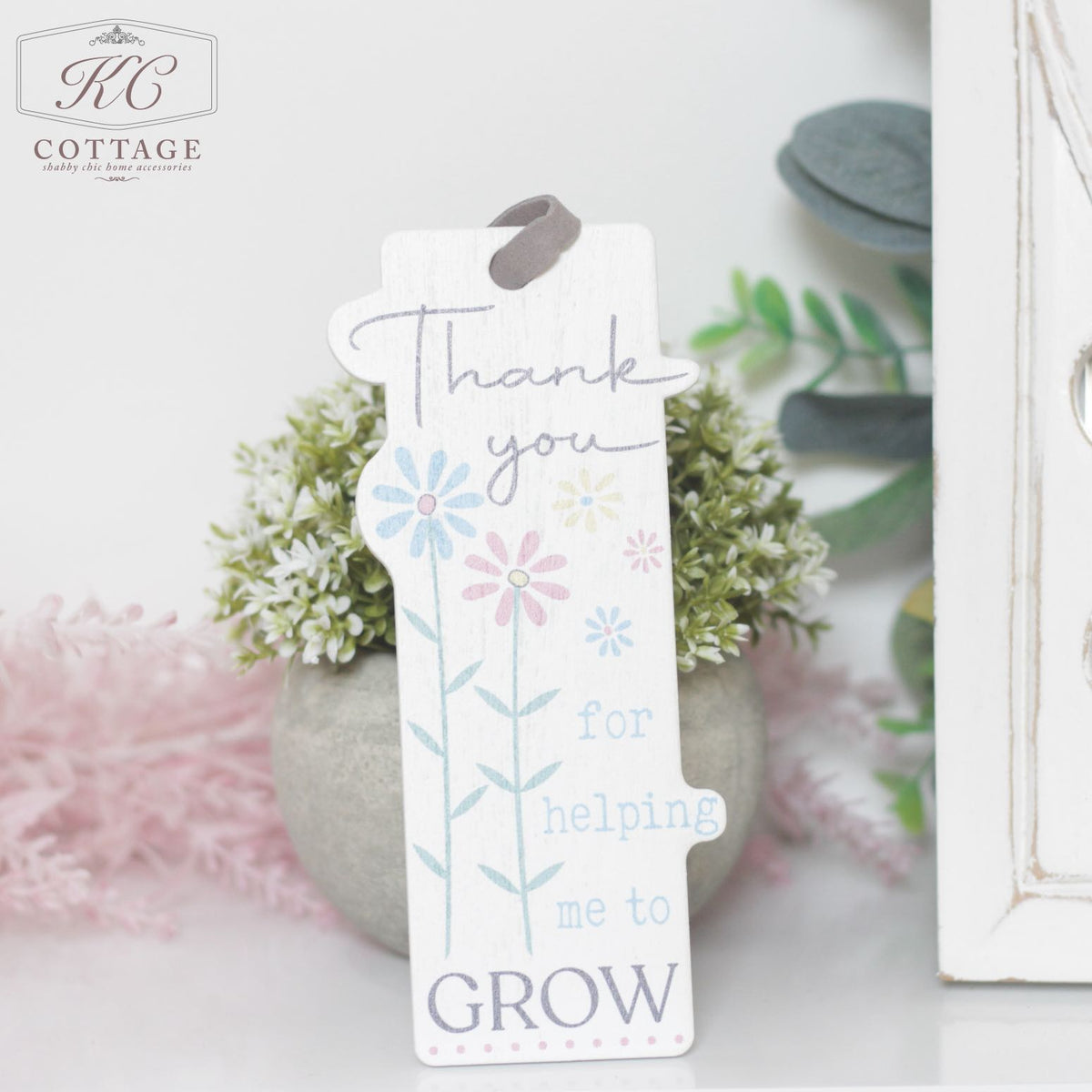 A wooden Teacher Gift - Thank You Bookmark stands upright in a plant pot. The bookmark features pastel-colored flowers and the text "Thank you for helping me to grow." Greenery surrounds the background, making it a charming piece of home decor.