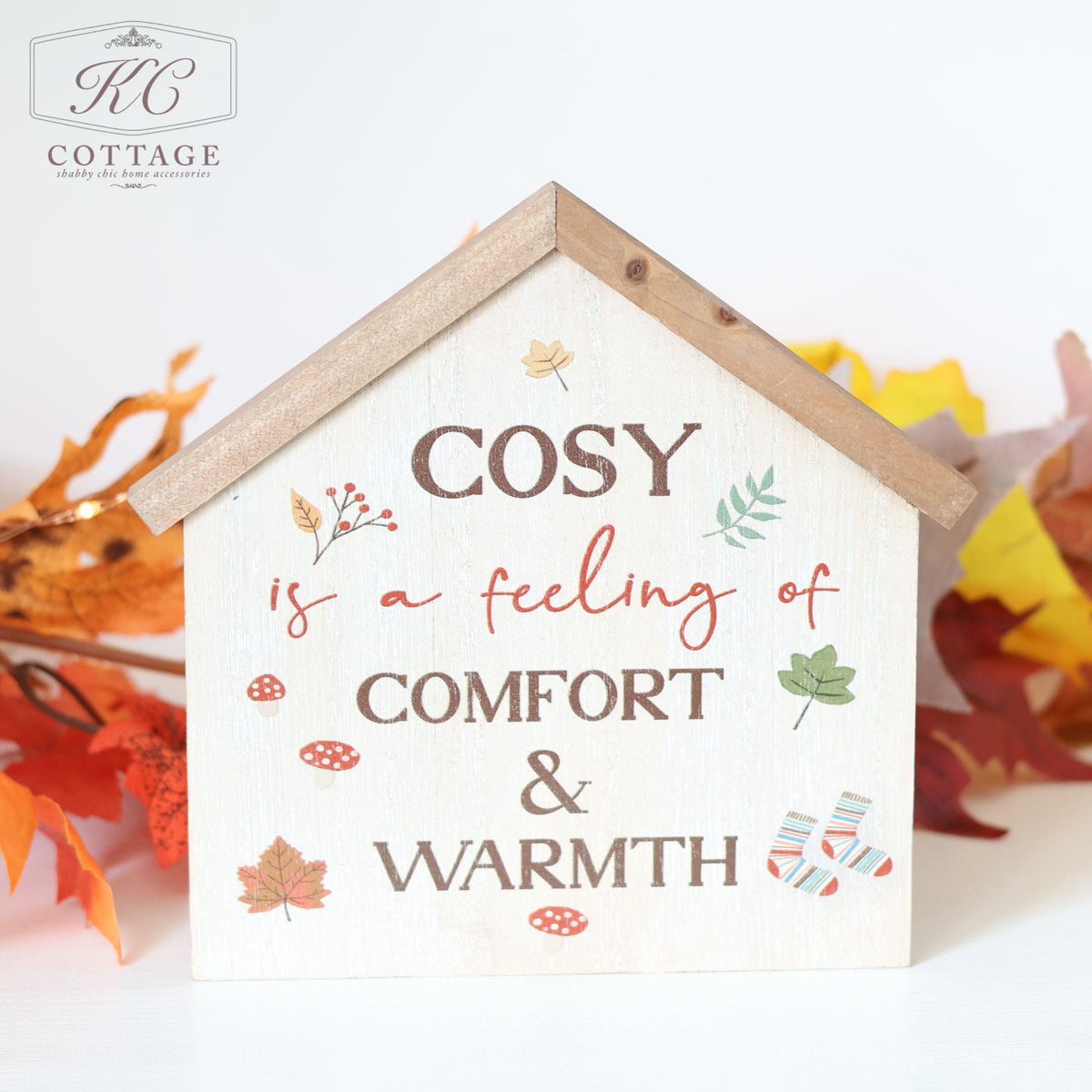 The Autumn Cosy Fall House Shaped Sign is a decorative wooden sign in the shape of a house, featuring the text "Cosy is a feeling of comfort & warmth," surrounded by illustrations of autumn leaves, acorns, mushrooms, and striped socks. The top corner displays the "KC Cottage" logo.