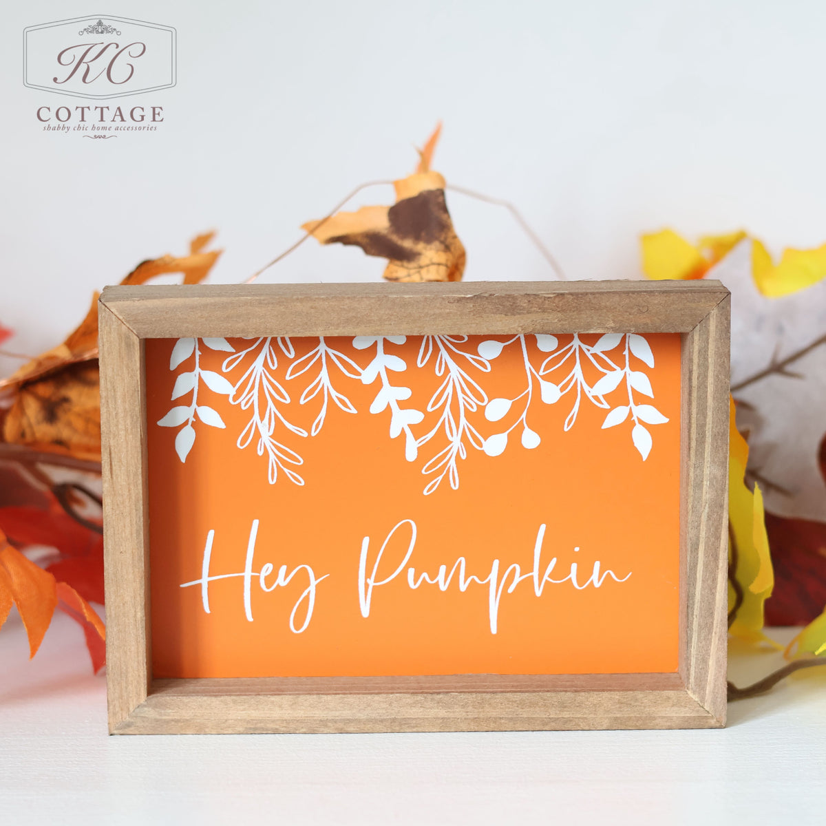 The Autumn Pumpkin Patch Sign, featuring a charming wooden frame and an orange background, proudly displays the phrase "Hey Pumpkin" in elegant white script. Delicate white leaf and vine silhouettes embellish the area above the text. This freestanding sign is showcased against a white wall with autumn leaves scattered around it, and the "KC Cottage" logo is prominently placed in the top left corner.