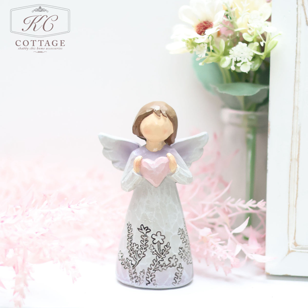 A Standing Angel Ornament with Love Heart holding a pink heart and wearing a long dress with intricate floral designs stands in front of a bouquet of white and pink flowers. The scene, perfect for home decor, is soft and pastel-colored.