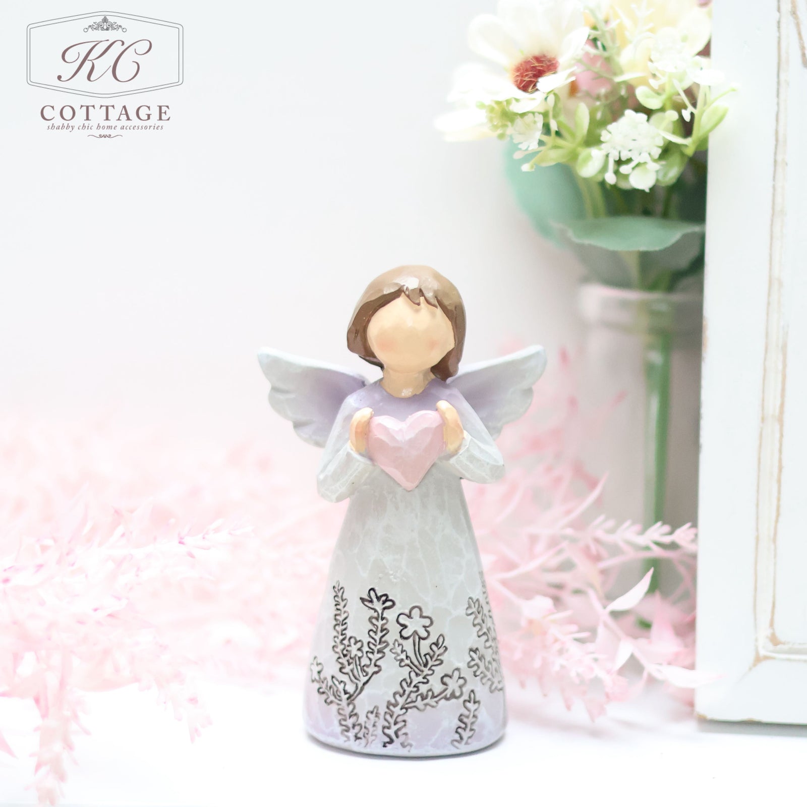 A Standing Angel Ornament with Love Heart holding a pink heart and wearing a long dress with intricate floral designs stands in front of a bouquet of white and pink flowers. The scene, perfect for home decor, is soft and pastel-colored.