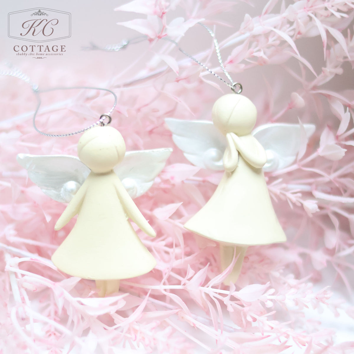 Two Large Hanging Angels with minimalist designs hang from silver strings amidst soft pink foliage. Perfect for home decor, both angels have smooth, featureless faces and small white wings. One angel has its hands clasped together, while the other has its hands at its sides.