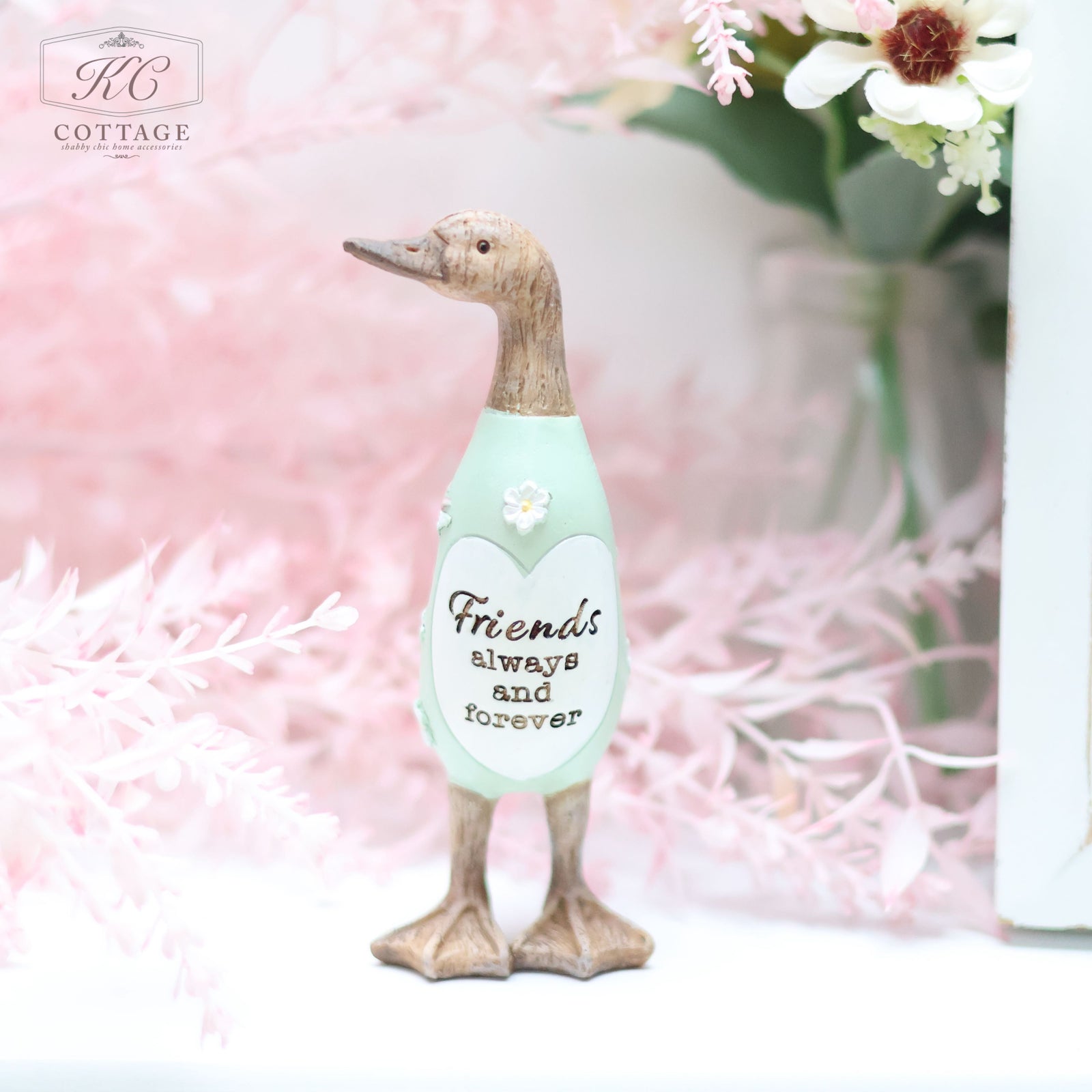 A Daisy Duck Ornament made of wood stands on a pastel-colored surface. The duck wears a mint-green outfit with a heart-shaped emblem that reads, “Friends always and forever.” Delicate pink and white flowers fill the background, enhancing the scene's charm, making it perfect for home decor.