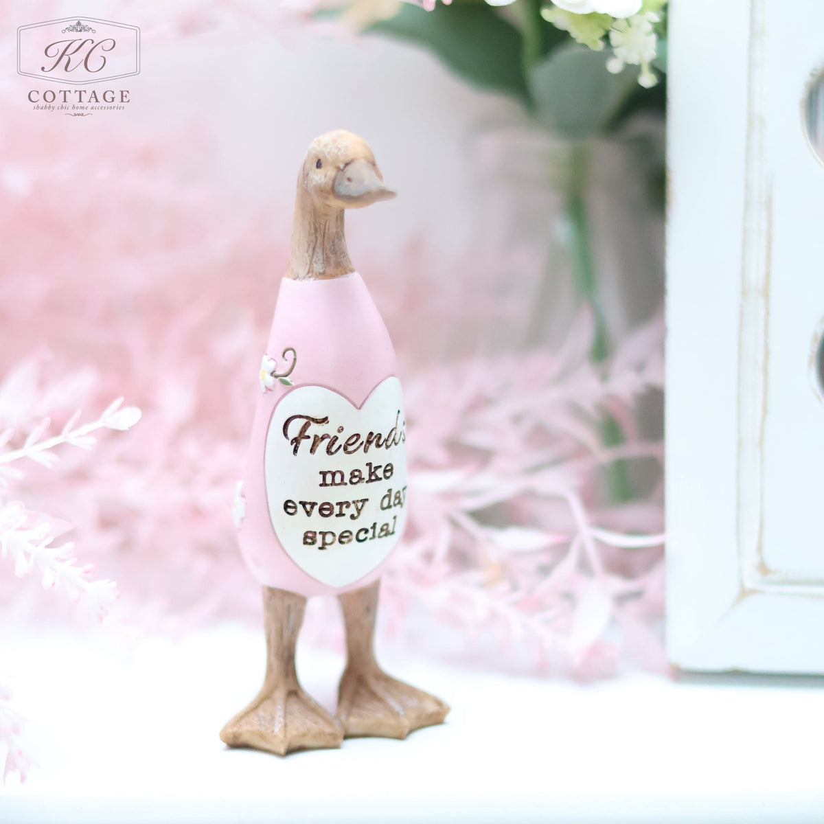A Daisy Duck Ornament wearing a pink dress with the text "Friends make every day special" stands on a white surface. Pink flowers and green leaves in the blurred background create a soft and serene atmosphere, making it an ideal piece for home decor.