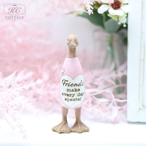 A small Daisy Duck Ornaments figurine wearing a pink outfit with a heart and the words "Friends make every day special" stands on a white surface. Pink flowers and greenery are blurred in the background, making it a charming addition to your home decor.