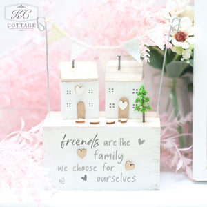 A Wooden Sentiment Houses with Bunting, adorned with small heart cut-outs and a tiny tree. It sits on a rectangular base with the inscription, "Friends are the family we choose for ourselves." Pink flowers are in the background, making it a charming piece of home decor.