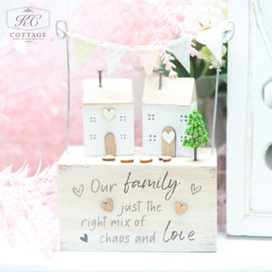 A charming home decor piece featuring two small houses, a tree, and a string of triangular flags. The white base showcases the engraved text, "Our family, just the right mix of chaos and love." The background includes soft pink foliage and part of a white shelf. This product is called Wooden Sentiment Houses with Bunting.