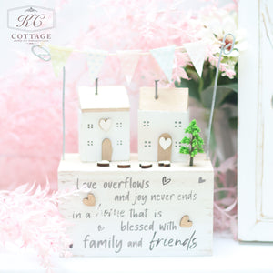 A charming piece of home decor featuring two small, white wooden houses with heart details, standing on a block with an inspirational quote about love, joy, and family. Pastel-colored bunting flags hang above the houses against a backdrop of pink foliage is the Wooden Sentiment Houses with Bunting.