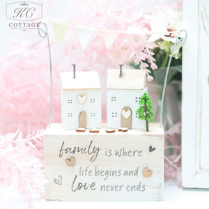A wooden sentiment houses with bunting block features two small white houses with heart-shaped windows and doors. A miniature tree stands beside them. The block is inscribed with the phrase, "Family is where life begins and love never ends," and is adorned with pastel bunting, making it a charming piece of home decor.