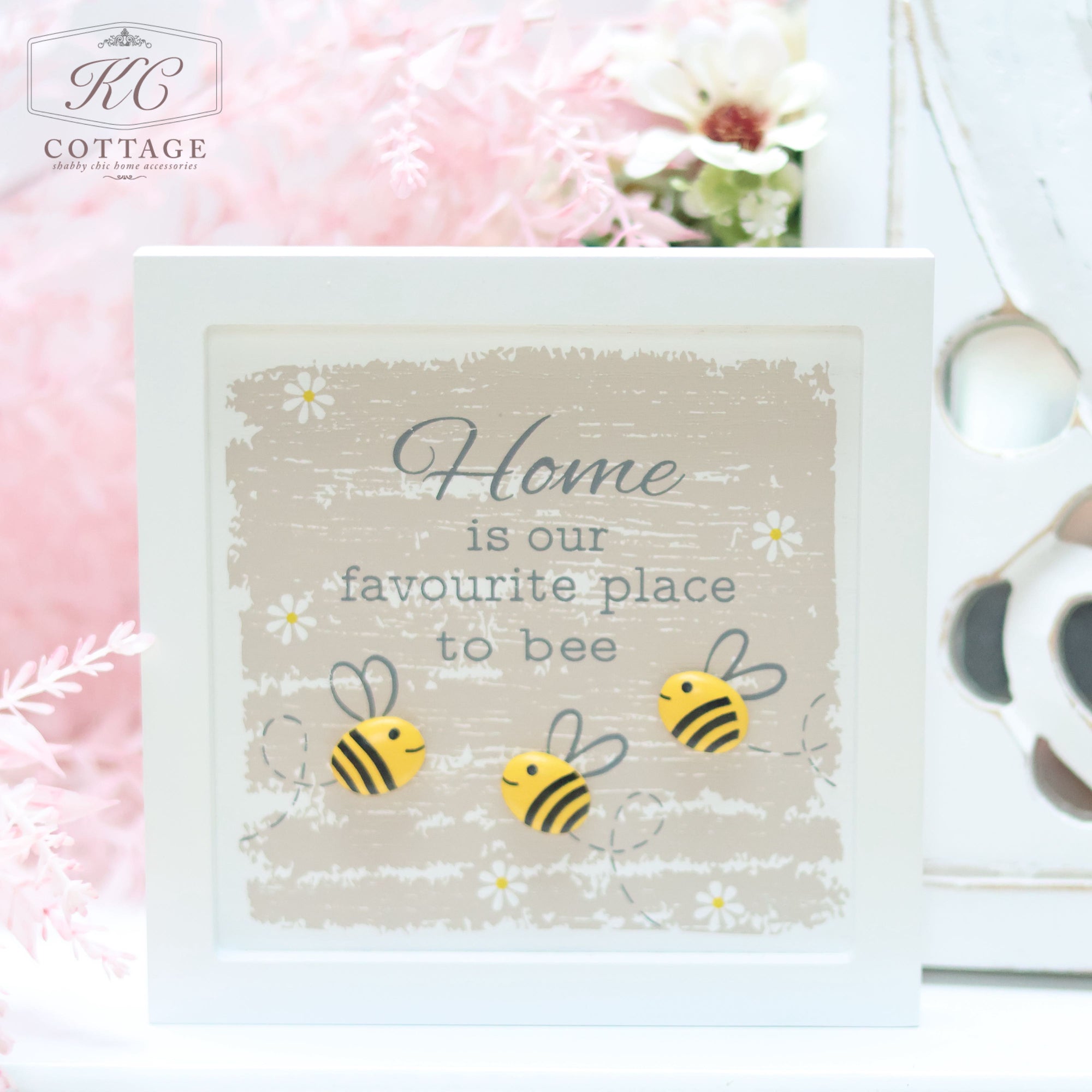 A framed decorative sign reads, "Home is our favourite place to bee," with illustrations of four bees flying around. The frame, an ideal piece of home decor, is adorned with a floral background and set against a softly focused pink backdrop with flowers.