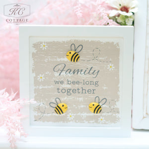 A square wooden Pebble Bee Sign with the sweet sentiment "Family we bee-long together" is placed in a soft, pastel-colored setting. The sign features illustrations of three bees and a rustic, weathered paint background. Pink flowers and light decor surround this charming piece of home decor.