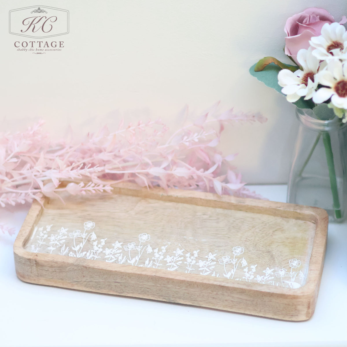 A Rustic Wooden Etched Tray with Flowers displayed on a light surface. The background features a pink feathery plant and a glass vase filled with white flowers and pink roses. The upper left corner has a "KC Cottage" logo.