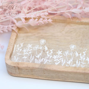 A Rustic Wooden Etched Tray with Flowers is positioned under soft pink artificial foliage. The tray has a charming, weathered appearance and is marked with a faint logo for KC Cottage in the top left corner.