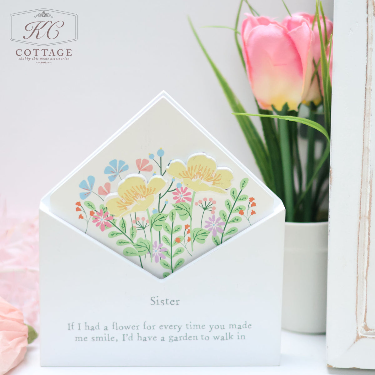 A decorative Wooden Floral Envelope Flower Box featuring colorful flowers is displayed partly enclosed in a white envelope. Labeled "Sister," it reads, "If I had a flower for every time you made me smile, I'd have a garden to walk in." Perfect for home decor, a pink tulip blooms softly in the background.