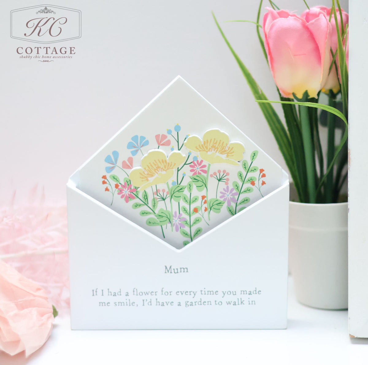 A Wooden Floral Envelope Flower Boxes with the label "KC Cottage" displays a white envelope adorned with colorful flowers. The base of the holder reads, "Mum, If I had a flower for every time you made me smile, I'd have a garden to walk in." This charming piece of home decor is complemented by a vase of pink tulips in the background.