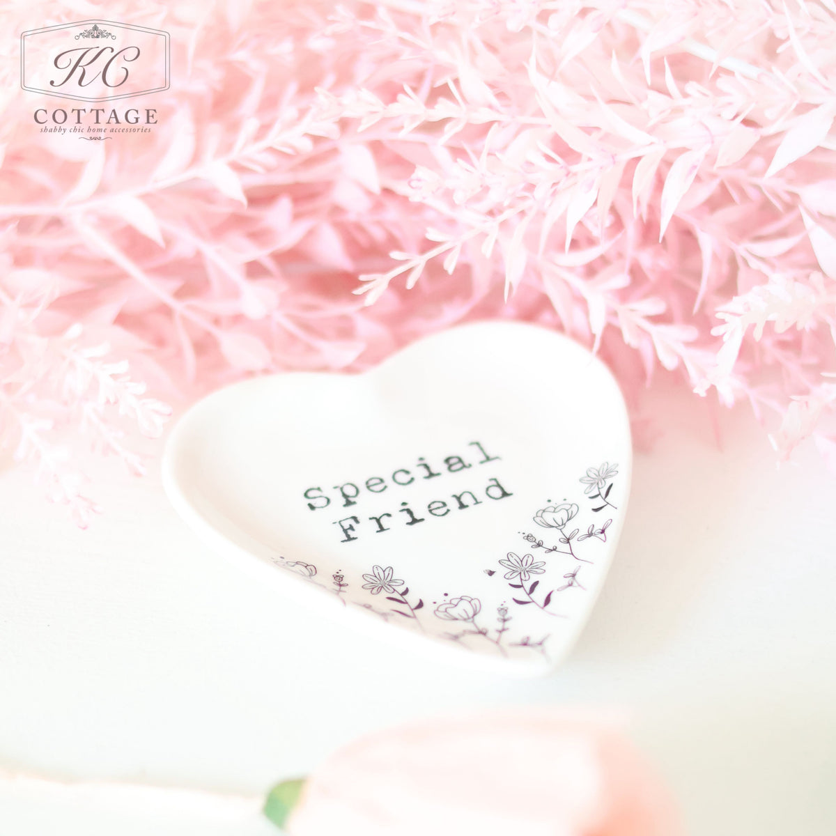 A small heart-shaped dish with the words "Gift Set - Best Friend" inscribed on it, along with a floral design at the bottom. The dish is placed on a light surface surrounded by soft pink, fern-like artificial flowers, making it a charming piece of home decor.