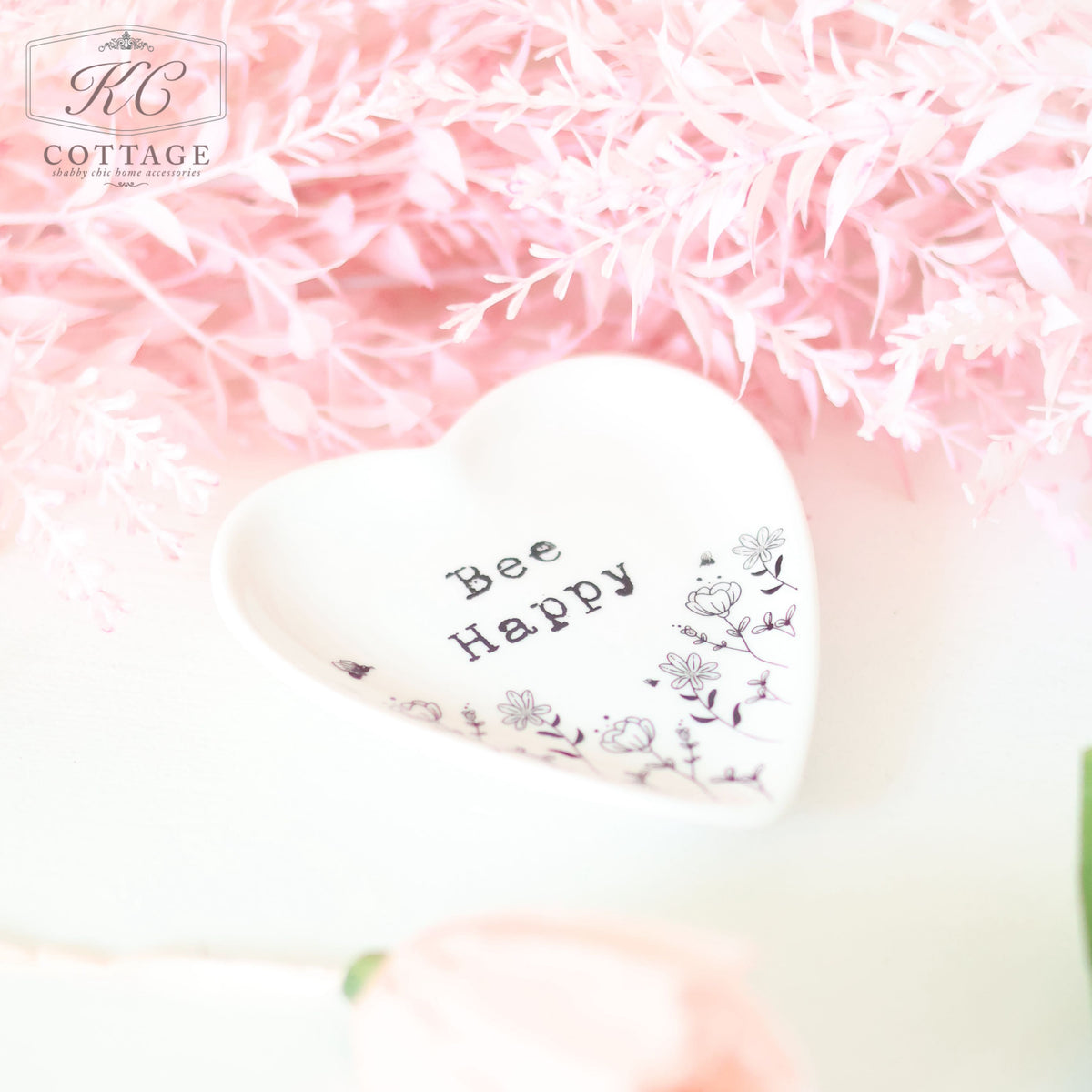 A heart-shaped dish with the words "Bee Happy" and floral illustrations sits on a white surface. Surrounding the dish are pink, feathery artificial flowers, adding charm to your home decor. A single pink rose is also visible in the bottom left corner of the Gift Set - Thinking Of You.