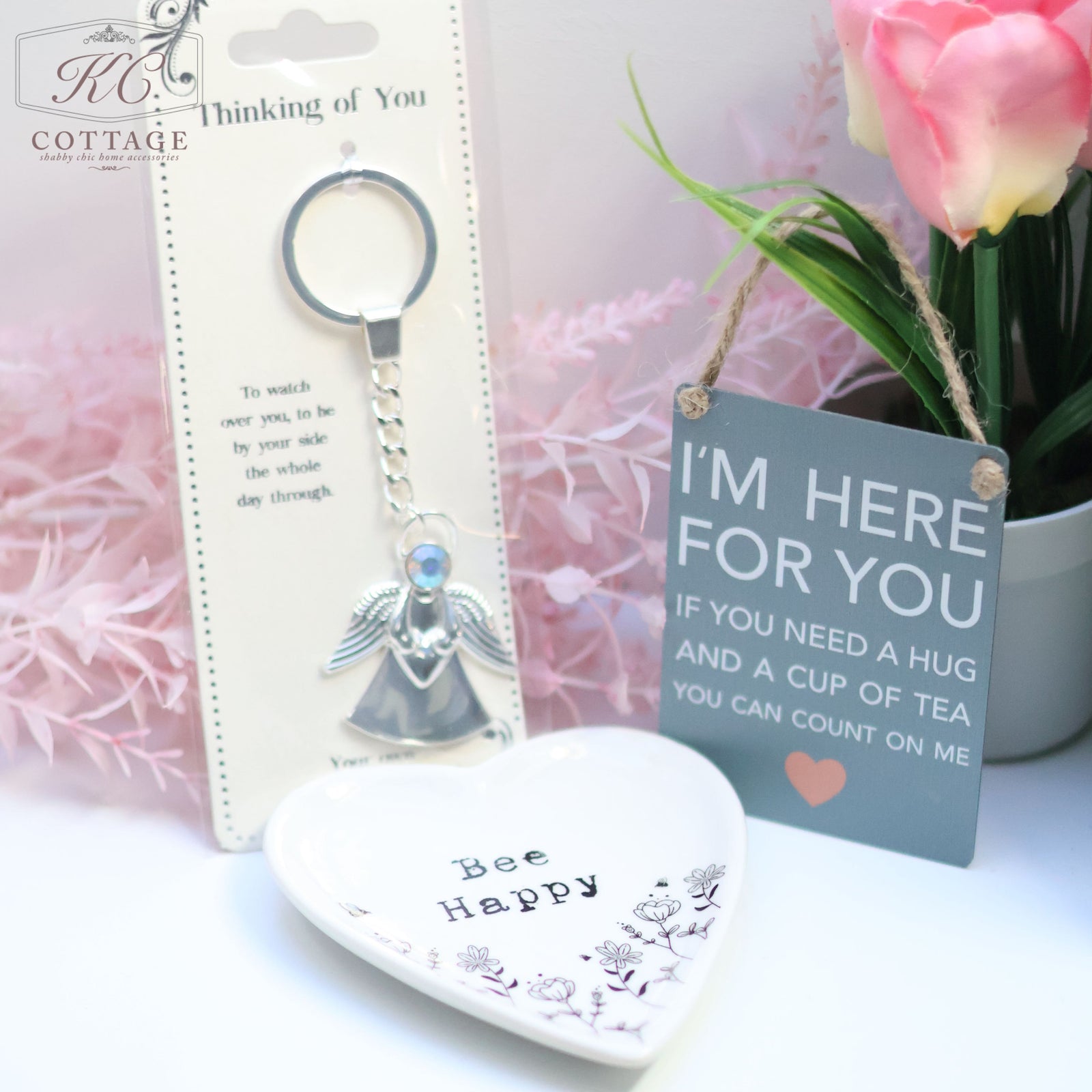 A gift set featuring a keychain with an angel charm, a heart-shaped dish with "Bee Happy" written on it, and a small sign that reads, "I'M HERE FOR YOU If you need a hug and a cup of tea you can count on me." Perfect for adding charm to your home decor, with a bouquet of flowers in the background. Introducing the Gift Set - Thinking Of You.