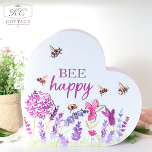 A Bees & Lavender Heart Plaque adorned with colorful illustrations of flowers and buzzing bees makes for charming home decor. The text on the plaque reads "BEE happy" in purple letters. The background features a soft-focus indoor setting with a potted plant in the distance.
