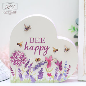 A heart-shaped decorative sign with the text "BEE happy" in purple and pink letters. It features illustrations of bees, flowers, and plants in varying shades of purple, pink, and green. Perfect for adding a cheerful touch to your home decor, the Bees & Lavender Heart Plaque is in the top left corner.