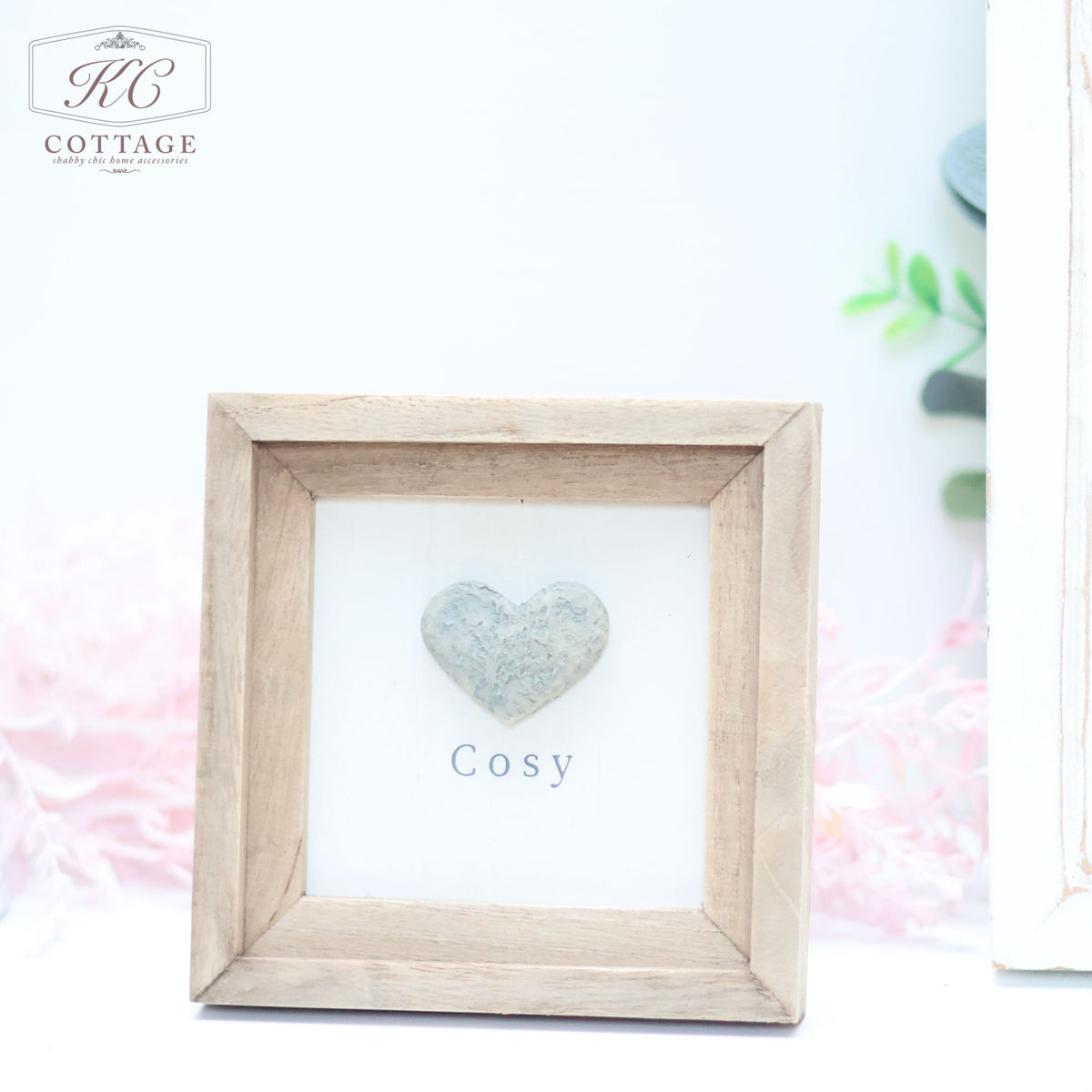A wooden picture frame with a rustic finish, containing a gray heart-shaped decoration and the word "Cosy" written below it. Decorative leaves and a pink feather are visible in the blurred background, adding to the charm of this home decor piece. The Pebble Heart Sign bears the logo "KC Cottage" in the top left.