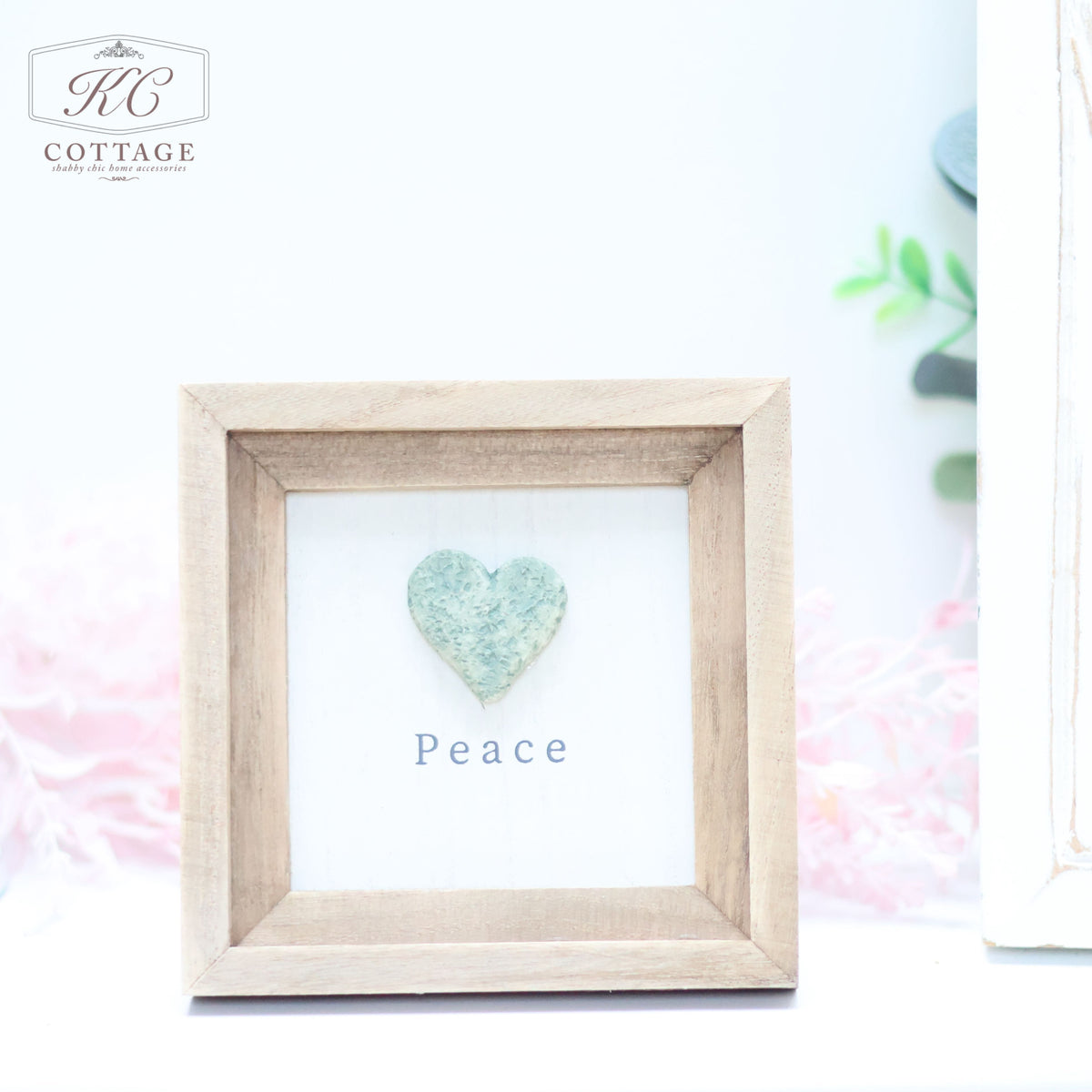 A small framed art piece with a beige wooden frame. Inside, a greenish-blue heart made of textured material is centered above the word "Peace" in simple black font. The background is a light, subtle shade, perfect for home decor. The logo "KC Cottage" is at the top left corner of the Pebble Heart Sign.