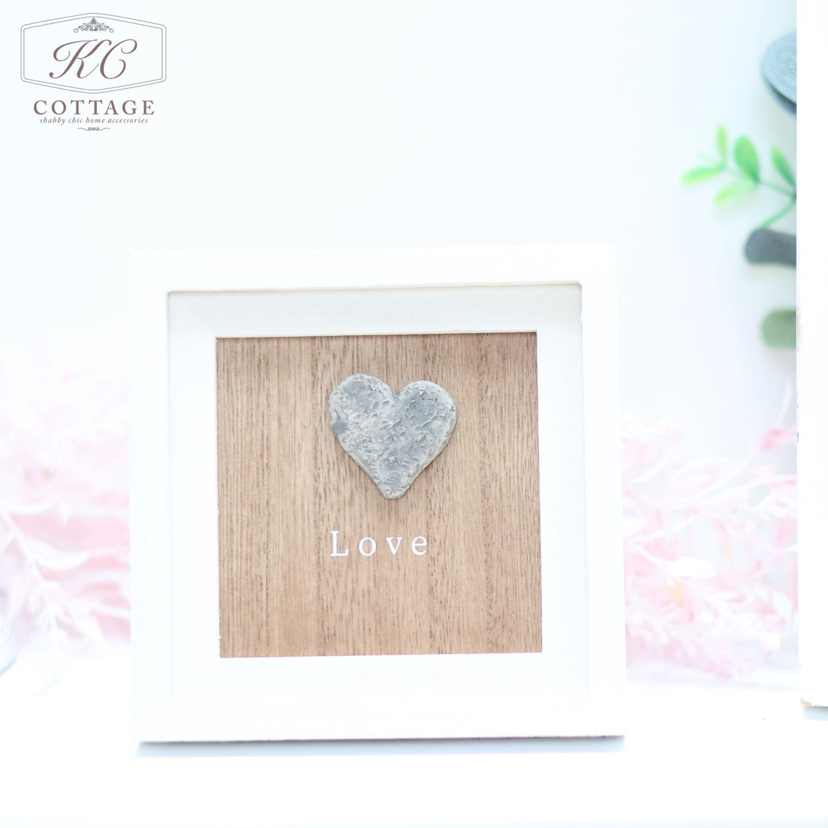 A framed decoration from KC Cottage featuring a wooden background with a raised heart shape and the word "Love" written in white beneath it. This charming Pebble Heart Sign has a white frame with soft pink flowers and green foliage softly blurred in the background.