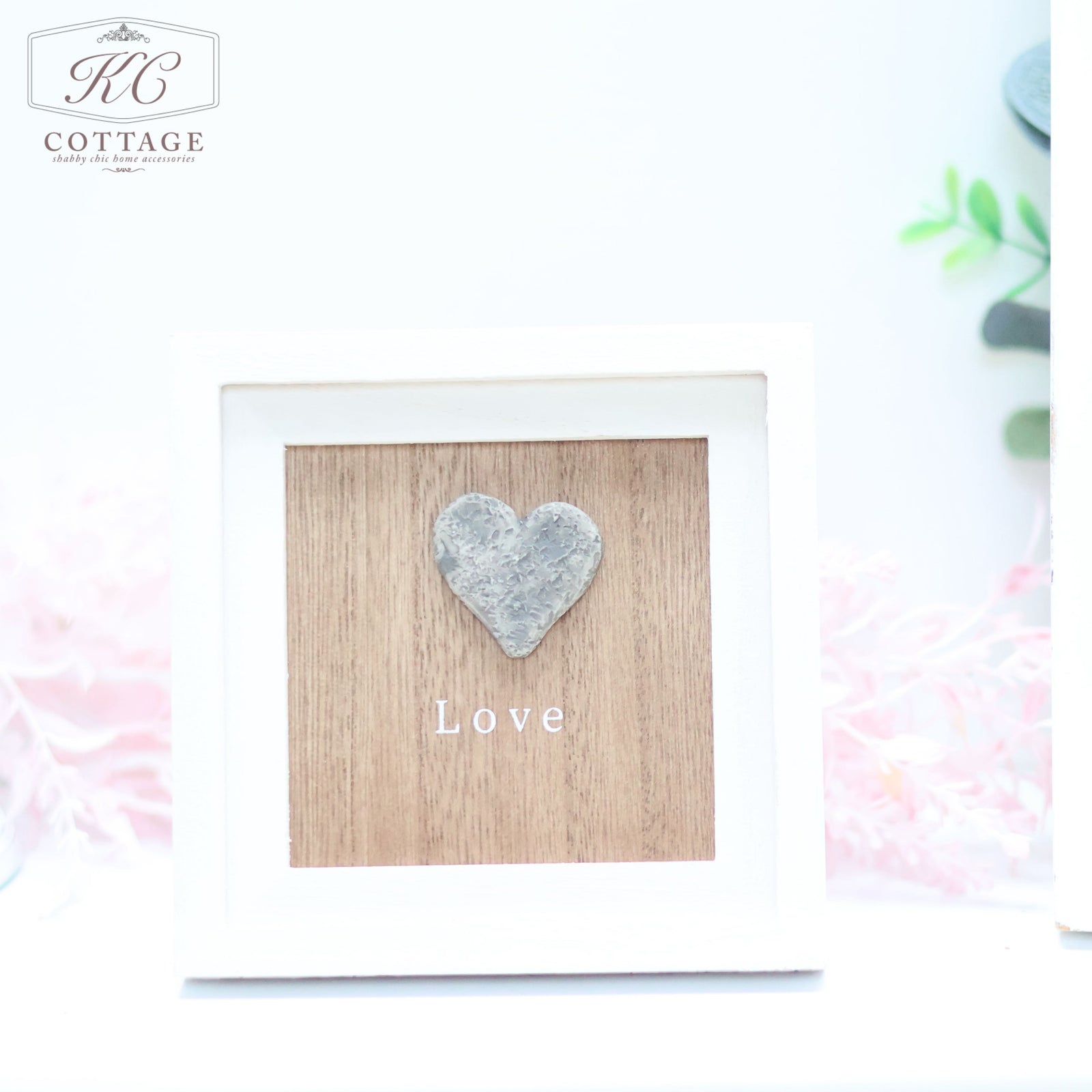 A framed decoration from KC Cottage featuring a wooden background with a raised heart shape and the word "Love" written in white beneath it. This charming Pebble Heart Sign has a white frame with soft pink flowers and green foliage softly blurred in the background.